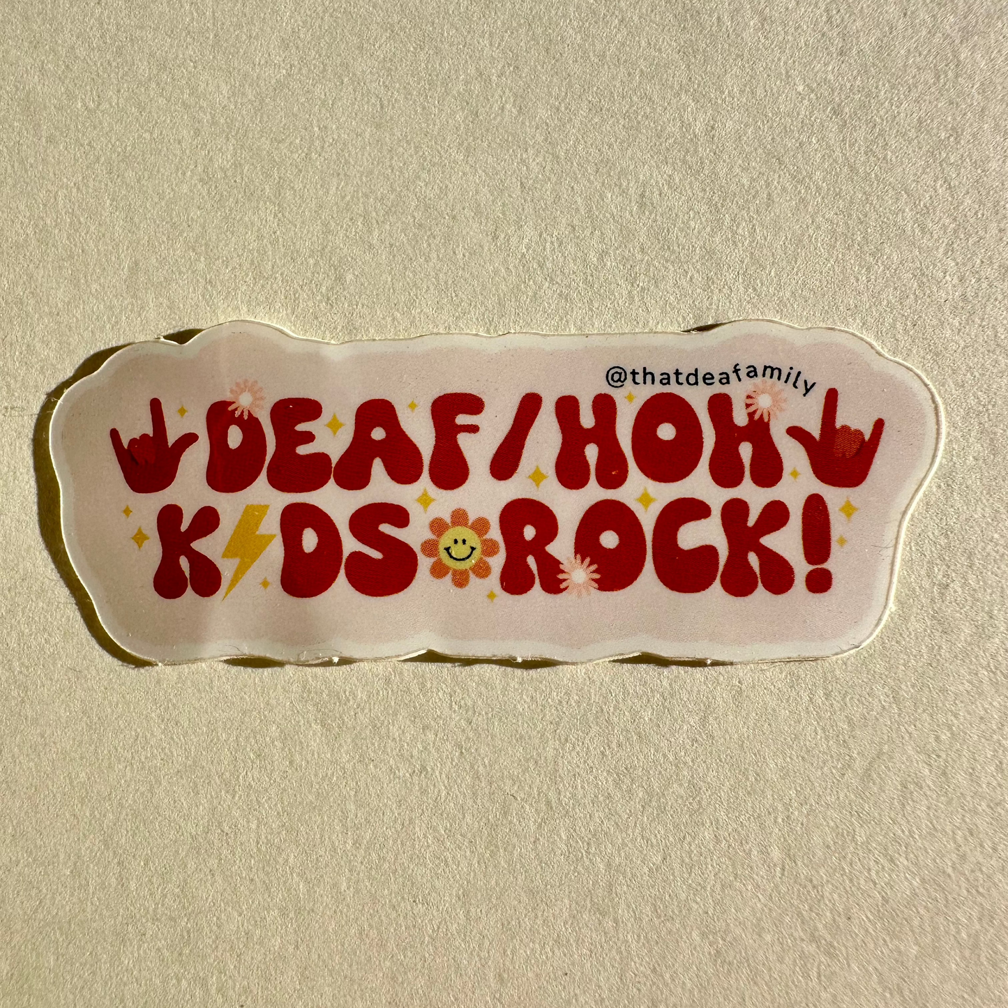 Clear vinyl sticker by @thatdeafamily featuring bold red text DEAF/HOH KIDS ROCK! with ASL I love you hand signs, yellow lightning bolts, and a smiling daisy on a textured tan surface